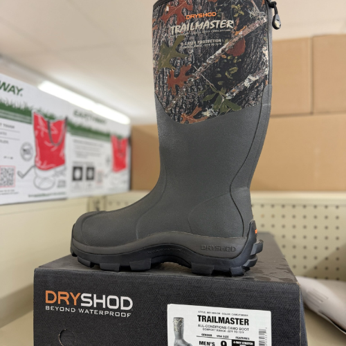 Gray waterproof boot with camouflage design on top, displayed on a box labeled "DRYSHOD Trailmaster." Store shelving and product signs in background.