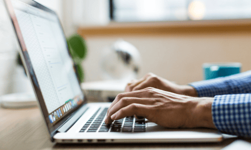 Hands typing on a laptop keyboard, with a blurred window and lamp in the background. The scene conveys focus and productivity in a workspace.