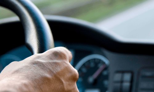 Close-up of a person's hand gripping a steering wheel, with blurred dashboard and road. The scene conveys focus and movement, suggesting driving.