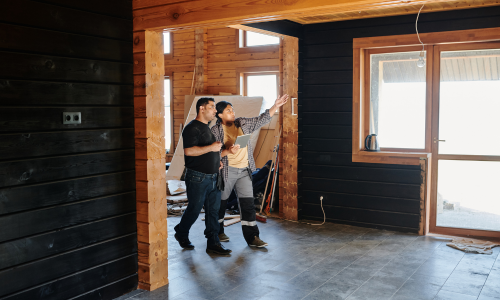 A couple stands in a partially renovated wooden room with large windows, examining the space. The atmosphere is hopeful and anticipatory.