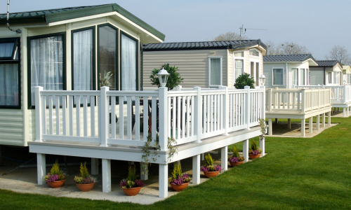A row of tidy mobile homes with white-railed porches, potted flowers, and green lawns under a bright, calm sky.