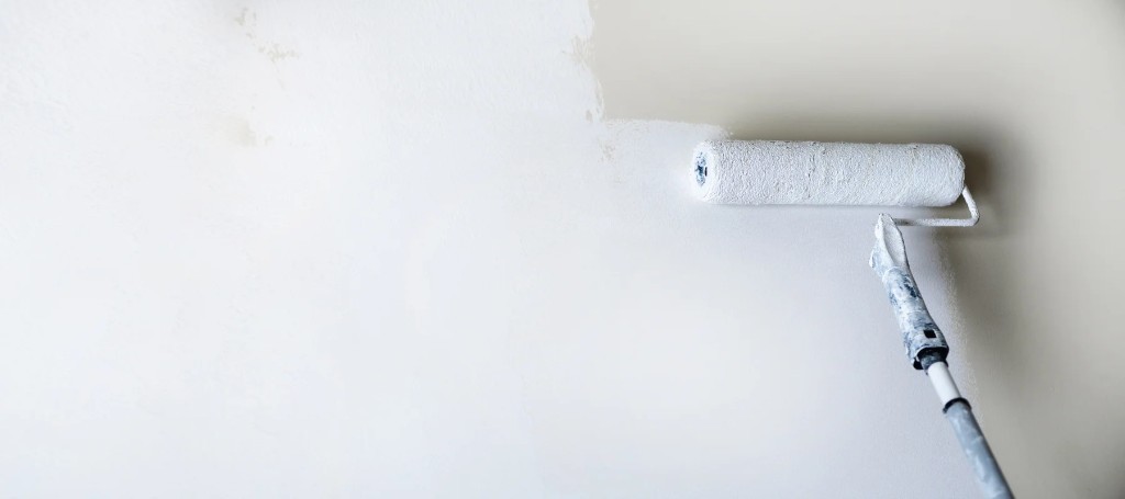 A paint roller applies fresh white paint to a wall, creating a clean, minimalist surface in a calm, neutral setting.