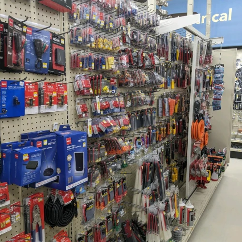 Hardware store aisle packed with cables, adapters, electronics, tools, and accessories on pegboards, creating a busy, well-organized retail scene.