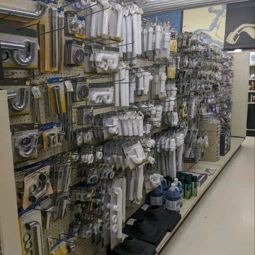 Hardware store aisle displaying rows of plumbing supplies—pipes, valves, fittings, hoses, and tools—organized on pegboard shelves.