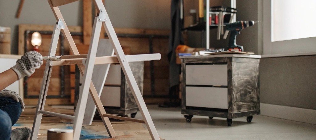 Person painting in a workshop beside an A-frame ladder, with a rolling tool cabinet, supplies, and bright window creating an industrious mood.