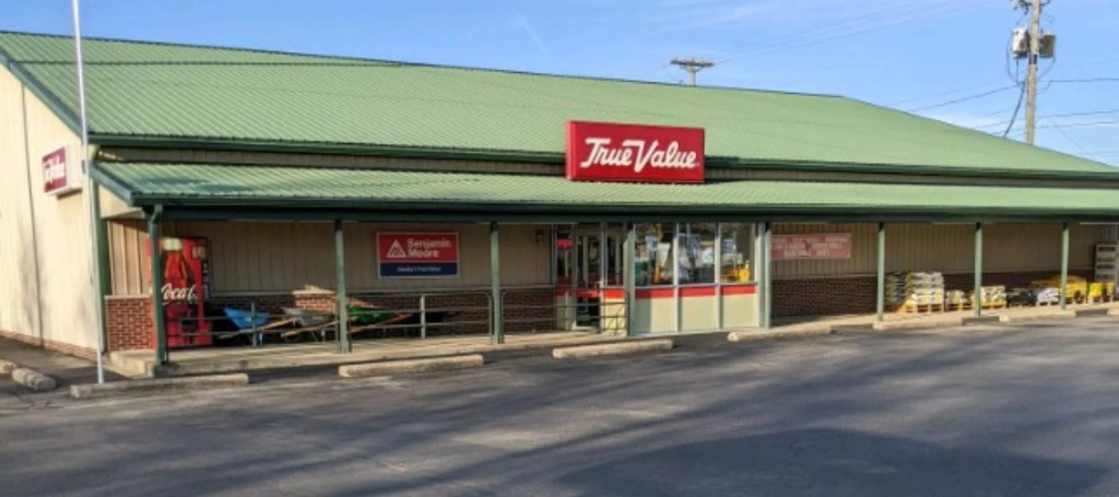 Exterior of a True Value hardware store with a green roof, red sign, storefront windows, carts, and an empty parking lot in daylight.