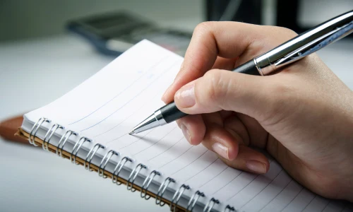 Close-up of a hand holding a black-and-silver pen, writing in a lined spiral notebook on a desk in an office; focused, productive mood.