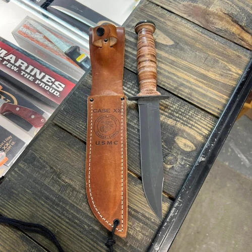 Vintage military-style combat knife with ribbed wooden handle beside a brown leather USMC sheath, displayed on a weathered wooden table.