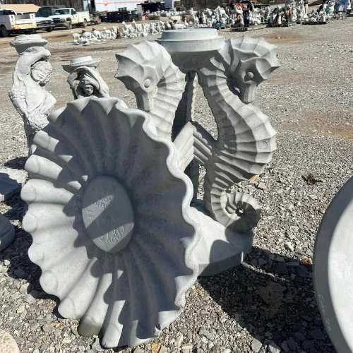 Gray stone sculpture of three seahorses emerging from scalloped shells, displayed outdoors in a gravel market under bright sunlight.