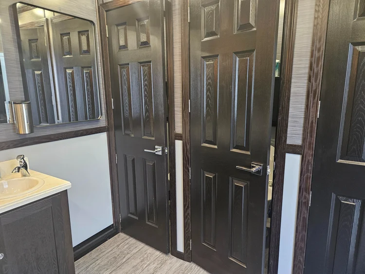 A dim, modern bathroom seen through dark glass partitions, with a white toilet, wall-mounted sink, shower fixtures, and a lit mirror; the mood is quiet and subdued.