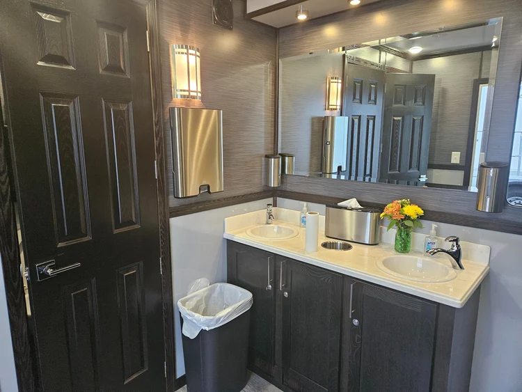 A compact modern bathroom with dark cabinets, a wood floor, white sink basins, and a large mirror; soft lighting creates a clean, calm feel.