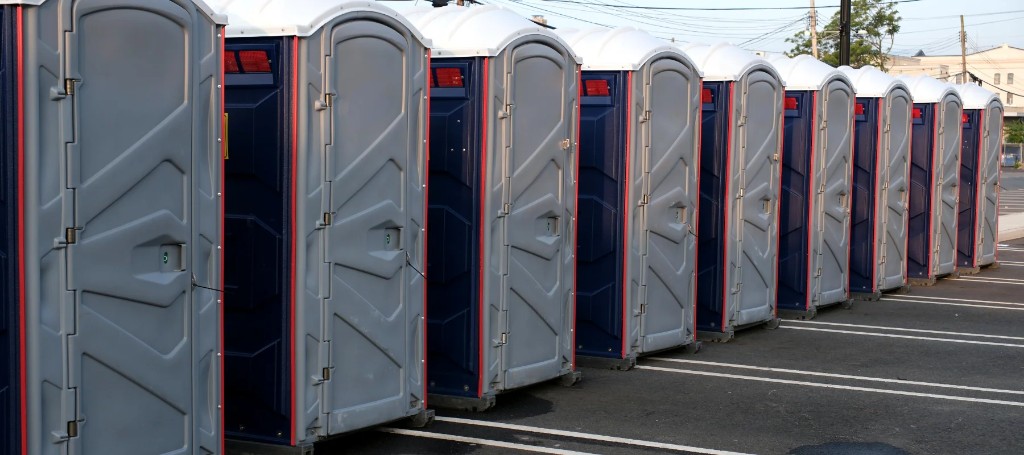 A long row of blue portable toilet booths stands in a parking lot, doors facing the viewer and shrinking into the distance under a bright sky.