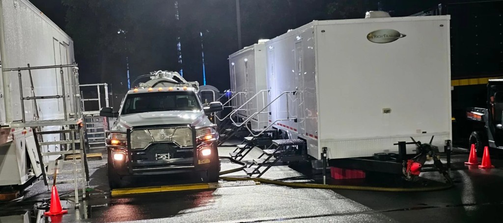 A dark, cinematic loading dock with a black delivery truck facing forward beside a white box trailer, cones and equipment on wet pavement.