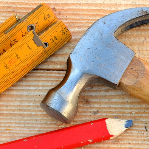 A steel hatchet, yellow folding ruler, and red pencil rest on a weathered wooden surface, evoking hands-on carpentry and practical workshop work.