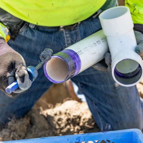 A worker in a neon yellow safety shirt and gloves holds white PVC pipe fittings with purple primer over a trench, preparing plumbing connections.