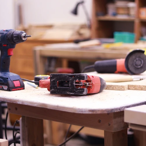 Cordless drill and red angle grinder rest on a worn workbench in a woodworking shop, with tools and shelves blurred behind them, creating a practical workshop mood.
