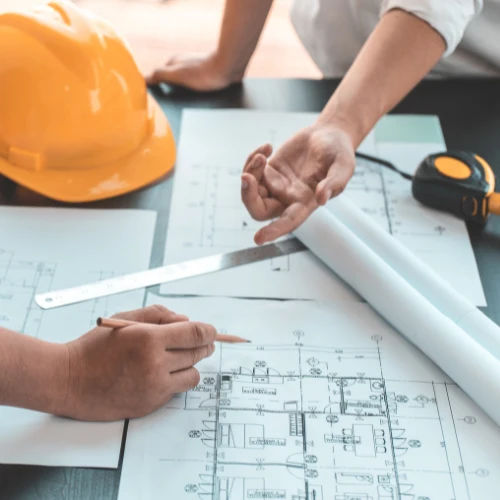 An architect points to a building blueprint as a colleague marks measurements with a pencil; an orange hard hat and tape measure suggest focused teamwork on a construction plan.
