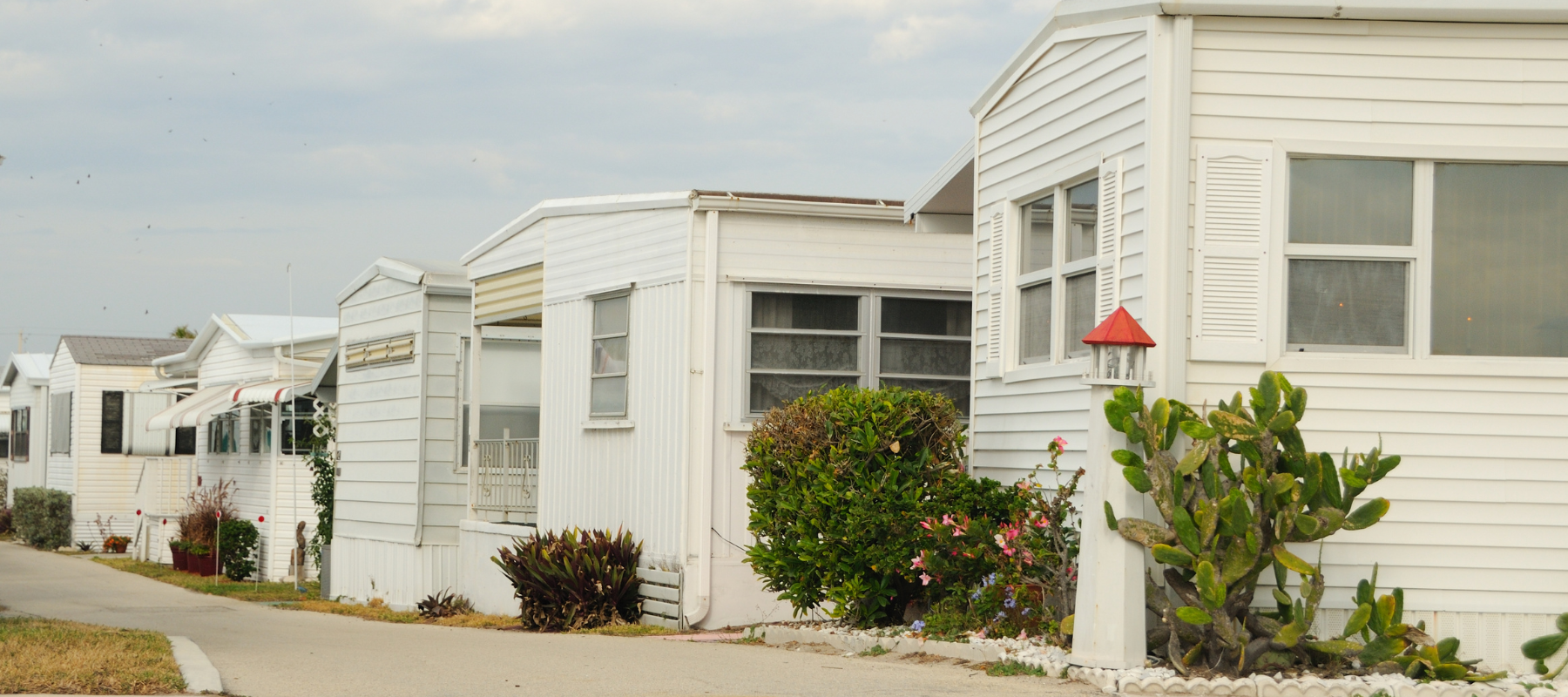 Row of white mobile homes with small gardens and shrubs, under a cloudy sky. The scene is calm and orderly, with a paved path in the foreground.