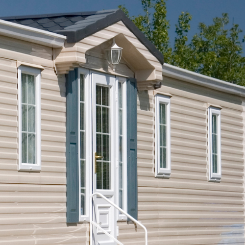 A neat beige-sided home exterior features a white glass-paneled front door, blue-gray shutters, slim windows, and a small white stair rail beneath a sunny sky.