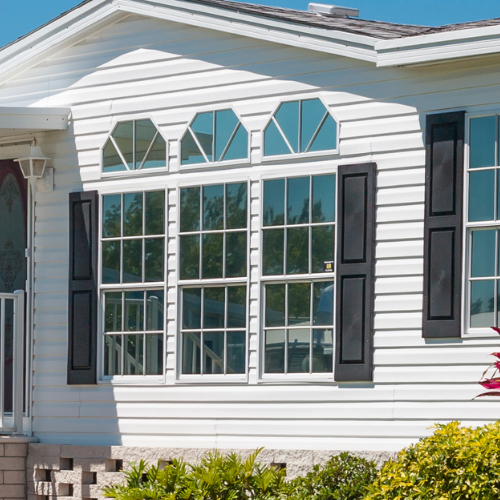 Sunlit white clapboard house with tall multi-pane windows, teal glass transoms, and black shutters, framed by garden greenery beneath a clear blue sky.