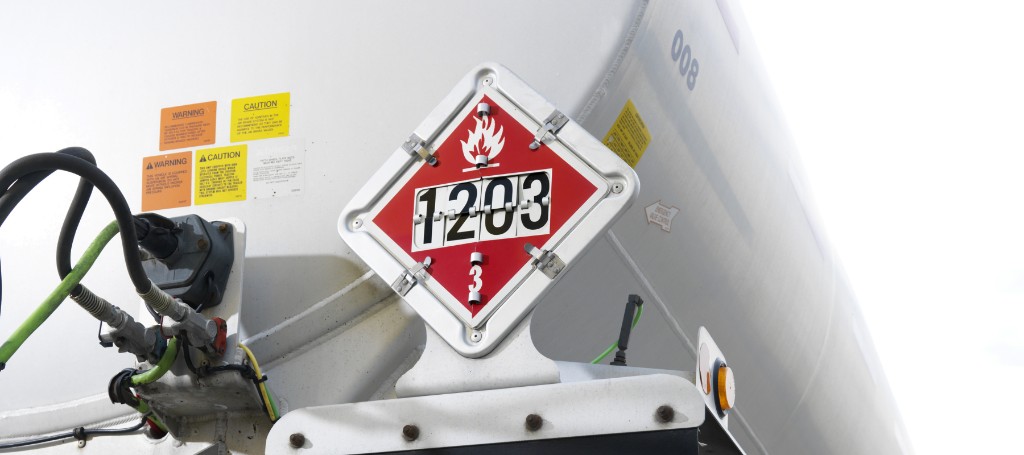 A red flammable-liquid placard marked 1203 mounted on a white tanker truck, with hoses and warning labels nearby.